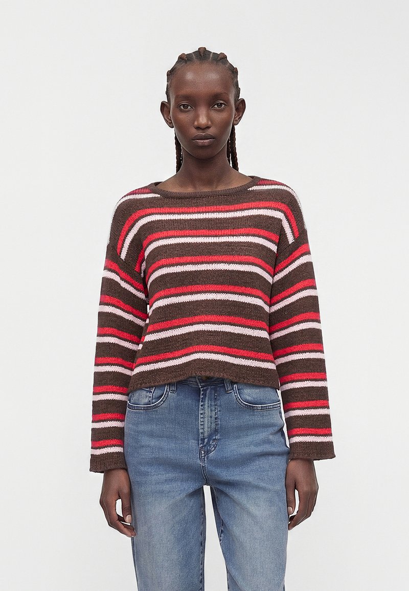Femme aux cheveux tressés portant un pull rayé horizontalement marron, rouge et rose et un jean bleu, debout devant un fond uni.