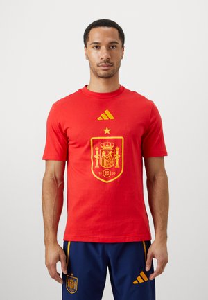 T-shirt en coton rouge avec un écusson de l'équipe nationale d'Espagne en jaune et le logo à trois bandes sur le col, à manches courtes. Design décontracté.