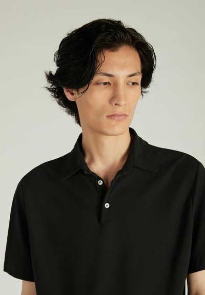 Polo negro hecho de algodón; presenta un cuello con botones y mangas cortas. Textura suave con detalles de costura sutiles.