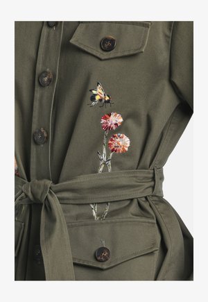 Olive green buttoned jacket with waist tie featuring embroidered bee and two pink and orange flowers near chest pocket.