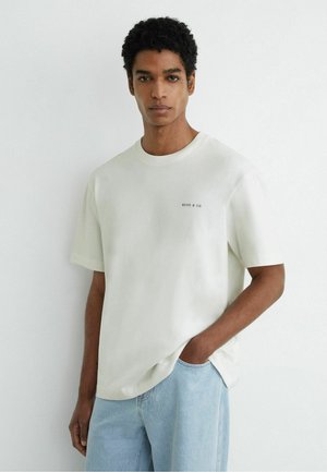 Jeune homme aux cheveux courts et bouclés, portant un t-shirt blanc Reiss & Co et un jean bleu clair, debout avec une main dans la poche.