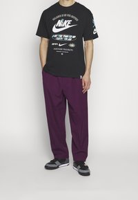 Černé bavlněné tričko s bílými a barevnými logy Nike, spárované s volnými fialovými kalhotami a černými teniskami.