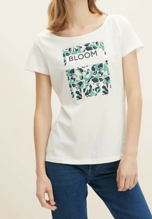 Kobieta w białym t-shircie z kwiatowym wzorem i słowem "BLOOM" napisanym wielkimi literami, w połączeniu z niebieskimi jeansami na beżowym tle.