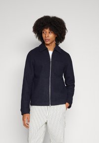 Navy wool zip jacket with a collar, featuring a front pocket, worn over white and navy striped pants. Smooth texture, casual design.
