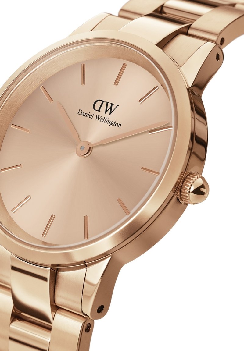 Montre en or rose avec un bracelet en métal poli, boîtier rond, cadran lisse et marqueurs d'heure minimalistes. Logo de la marque affiché sur le cadran.