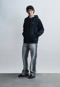 Jauns vīrietis stāv ar rokām kabatās, valkājot melnu džemperi ar kapuci, izbalējušas pelēkas džinsa bikses un melnus sporta apavus uz vienkārša fona.