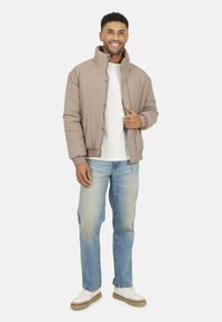 Giubbotto bomber imbottito color marrone chiaro con colletto alto e polsini elasticizzati, indossato sopra una camicia bianca e abbinato a jeans azzurri chiari e sneakers bianche.