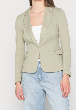 Frau trägt einen hellolivfarbenen Blazer mit Notch-Revers, Pattentaschen, Ein-Knopf-Verschluss, weißem Top und hellblauen High-Waist-Jeans.