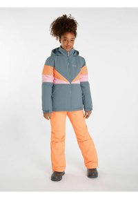Veste de ski colorée avec des motifs géométriques gris, rose et orange. Associée à un pantalon de ski orange vif et des bottes noires.