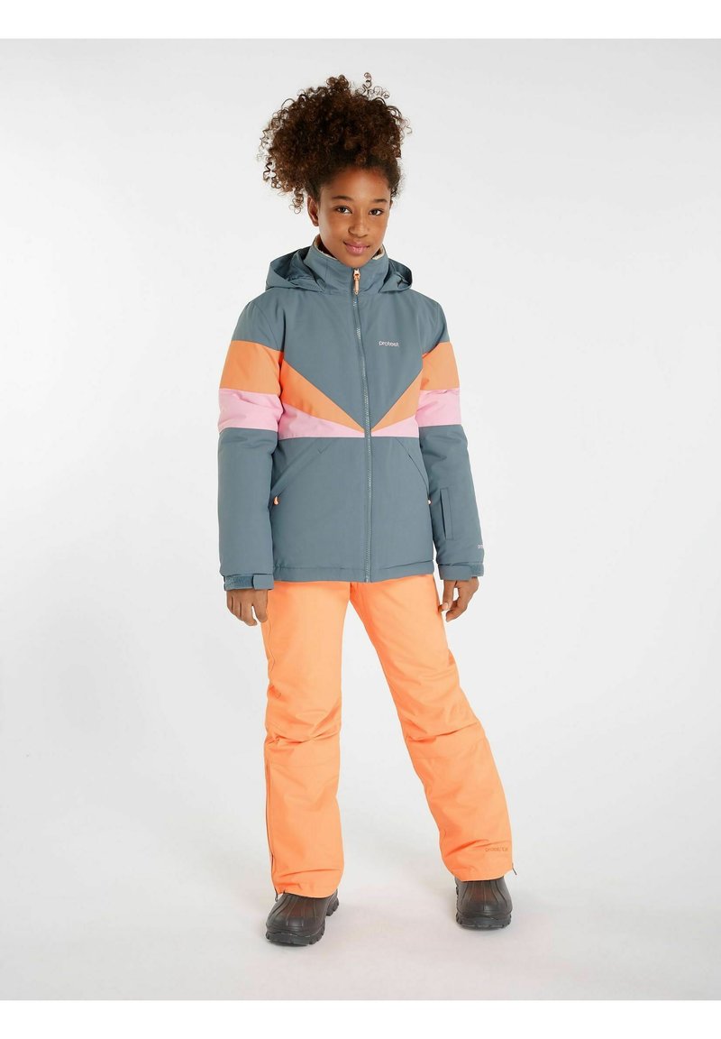 Veste de ski colorée avec des motifs géométriques gris, rose et orange. Associée à un pantalon de ski orange vif et des bottes noires.