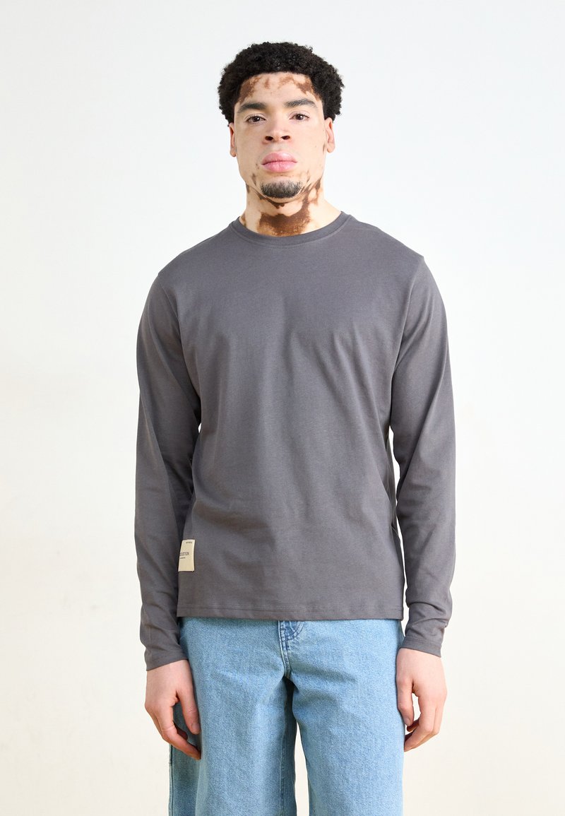Long-sleeve gray shirt made of cotton, featuring a crew neck and a small label on the lower side, paired with light blue denim jeans.