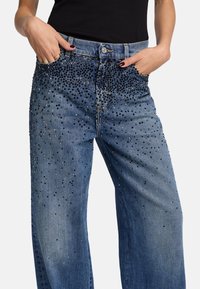 Hellblaue Jeans aus Denim mit weitem Bein, die über die Vorderseite verstreute schwarze und silberne Strasssteine und ein klassisches Fünf-Taschen-Design aufweisen.