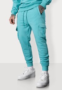 Türkise Jogginghose mit Seitentaschen, Kordelzugbund und gerippten Bündchen, kombiniert mit weißen Sneakers vor einem minimalistischen Hintergrund.