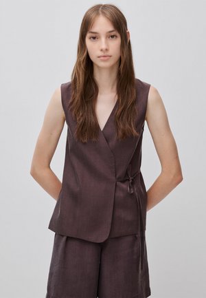 Femme aux longs cheveux bruns, portant un gilet cache-cœur sans manches marron foncé et un short assorti sur un fond gris uni.