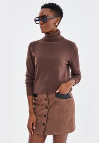 Brązowy sweter w serek zestawiony z brązową spódnicą z czarnymi kieszeniami i detalami w postaci guzików. Modelka nosi dużą parę okularów przeciwsłonecznych.