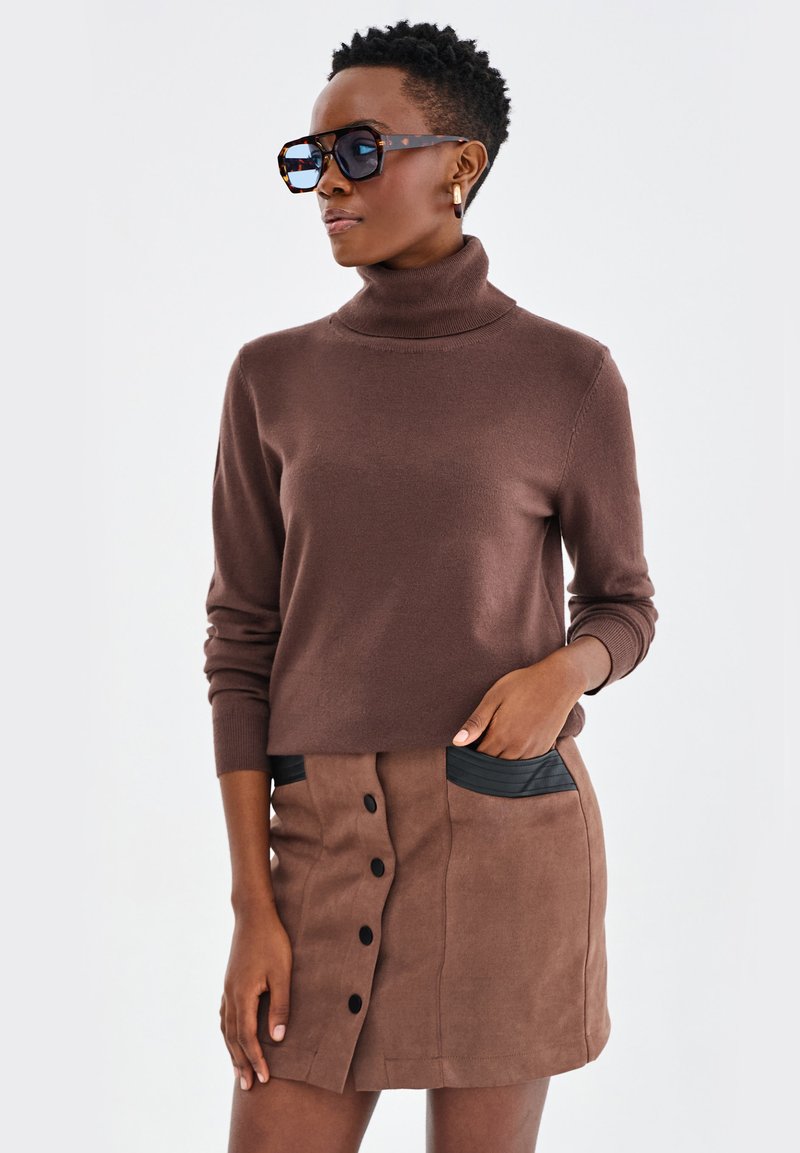 Brązowy sweter w serek zestawiony z brązową spódnicą z czarnymi kieszeniami i detalami w postaci guzików. Modelka nosi dużą parę okularów przeciwsłonecznych.