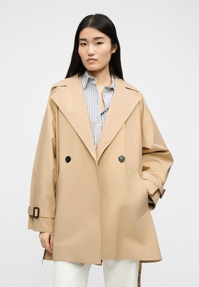 Γυναίκα με μακριά σκουρόχρωμα μαλλιά που φοράει μπεζ παλτό με διπλό πέτο πάνω από ριγέ πουκάμισο και λευκό παντελόνι, στέκεται μπροστά σε ένα απλό φόντο.