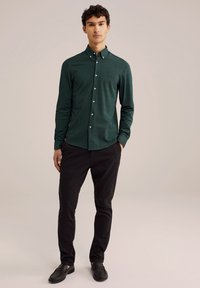 Temno zelena srajca s dolgimi rokavi, z ovratnikom in sprednjim zapiranjem na gumbe, subtilne teksture, v kombinaciji s črnimi ozkimi hlačami in črnimi čevlji.