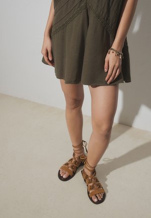 Frau trägt ein olivgrünes Kleid und braune Schnürsandalen, steht auf hellem Boden vor einer schlichten Wand, mit einem Charm-Armband am Handgelenk.