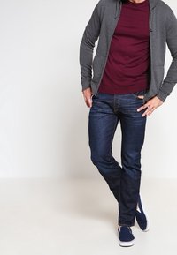 Grå zip-up hoodie över en vinröd skjorta, kombinerad med mörkblå jeans och marinblå slip-on sneakers. Enkel bakgrund, stående pose.