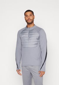 Grauer Langarm-Pullover mit Reißverschlusskragen, ausgestattet mit einem gepolsterten grauen Brustbereich und schwarzen Seitenakzenten. Nike-Logo auf der linken Seite.