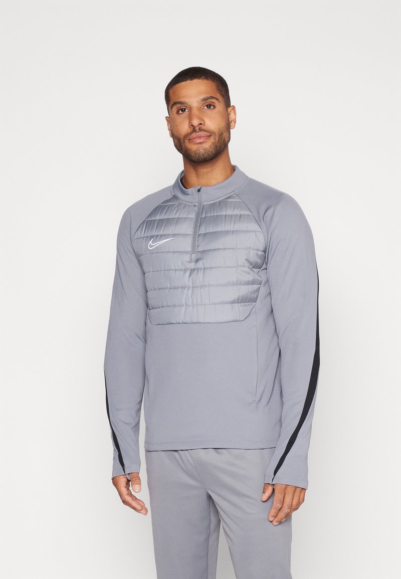 Grauer Langarm-Pullover mit Reißverschlusskragen, ausgestattet mit einem gepolsterten grauen Brustbereich und schwarzen Seitenakzenten. Nike-Logo auf der linken Seite.
