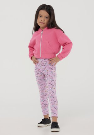 Cappotto corto rosa con zip e tasca frontale, abbinato a leggings rosa chiaro con un vivace motivo di unicorni. Sneaker nere completano il look.