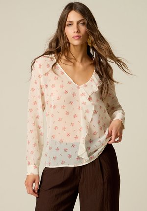 Frau mit langen braunen Haaren, die eine cremefarbene Bluse mit kleinen rosa Blumen und Rüschen trägt, kombiniert mit locker sitzenden dunkelbraunen Hosen.