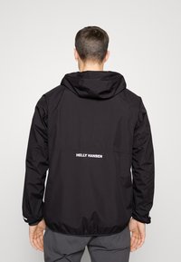 Schwarze wasserdichte Jacke mit Kapuze, die ein weißes Logo auf dem Rücken hat. Das Material scheint glatt zu sein, mit elastischen Bündchen und entspanntem Schnitt.