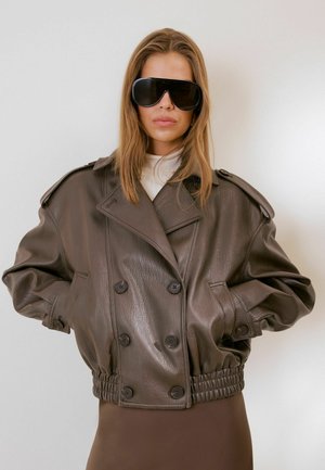 Femme portant une veste en cuir marron foncé oversize avec de gros boutons et des lunettes de soleil noires oversize, debout devant un fond clair uni.