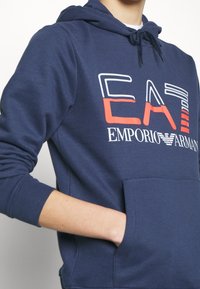 Mörkblå hoodie med framficka, dragsko på huvan och en iögonfallande vit/röd "EA7 EMPORIO ARMANI"-logotyp på bröstet. Slätt tyg i bomullsblandning.