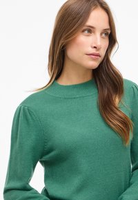 Grön stickad tröja med ribbad krage och långa ärmar. Har puffade axlar och en slät textur. Avslappnad passform och enkel design.