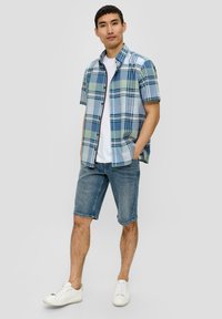 Korte mouwen geruite shirt in blauwe en groene tinten, gecombineerd met denim shorts. Model draagt witte sneakers. Casual, ontspannen pasvorm.