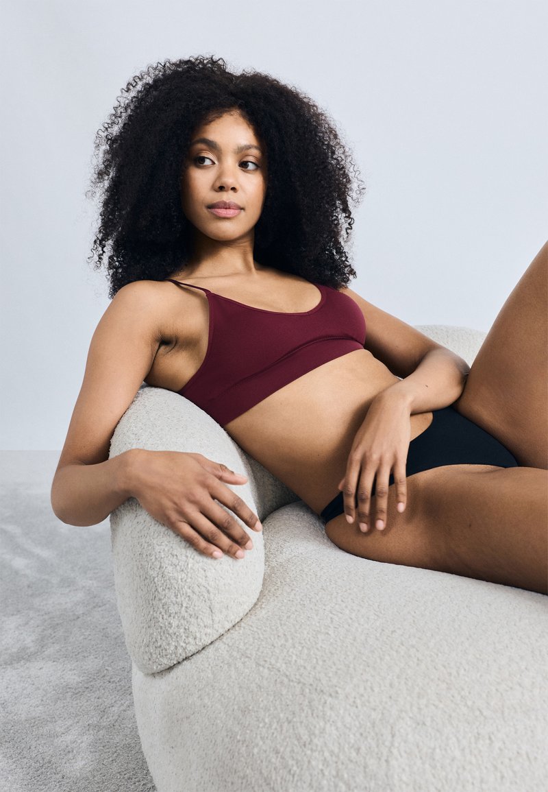 Sutiã desportivo mauve com alças finas; cuecas pretas de cintura alta. A modelo repousa numa sofá curvo texturizado de cor creme contra um fundo claro.