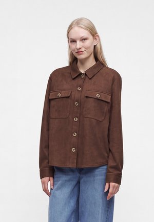 Ung kvinde med lyst, glat hår iført en brun jakke med knapper og to brystlommer samt blå jeans, stående foran en ensfarvet baggrund.