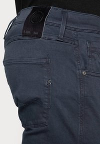 Navy blue jeans with a leather patch at the waist, metal accents, and a back pocket featuring a stitched pattern and a small logo.