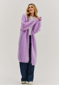 Dlhý kardigán s otvoreným predným dielom z huňatého, svetlofialového materiálu. Má široké rukávy a voľný strih, doplnený tmavými nohavicami a bielymi teniskami.