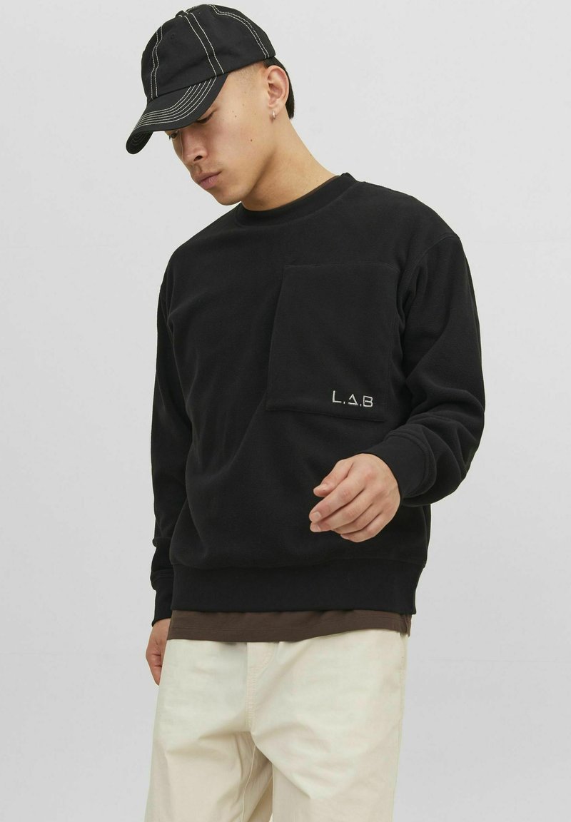 Čierny fleecový sveter s predným vreckom a logom "L.A.B.", nosený s čiernym šiltovkou. Svetlé nohavice dotvárajú celý vzhľad.