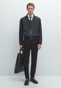 Zwarte ritssluiting jacket, witte shirt, zwarte stropdas en op maat gemaakte zwarte broek. Model houdt een zwarte leren tote bag vast en draagt zwarte enkellaarsjes.