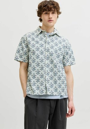 Jeune homme portant une chemise blanche à manches courtes avec des motifs floraux bleus sur un sous-vêtement gris et un pantalon plissé noir.