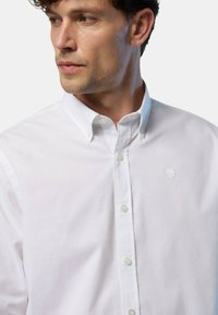 Camicetta bianca con bottoni in cotone, caratterizzata da un colletto classico, bottoni frontali e un piccolo logo sul lato sinistro del petto. Texture liscia.