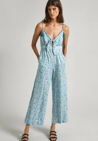 Blauwe jumpsuit met een patroon, voorzien van een strik vooraan, spaghettibandjes, brede benen en zakken. Accentueert met blauwe kwastjes op de bandjes.