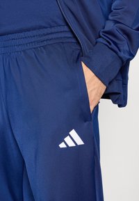 Mornarsko modri atletski hlače z teksturirano tkanino, s elastičnim pasom in belim Adidasovim logotipom na spodnjem delu noge.
