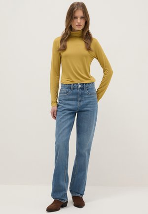 Vrouw met lang haar, gekleed in een mosterdgele coltrui, blauwe high-waisted jeans en bruine enkellaarsjes, staand tegen een effen achtergrond.