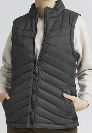 Personne portant une veste sans manches matelassée noire avec une fermeture éclair à l'avant et les mains dans les poches latérales, par-dessus une chemise beige clair à manches longues.