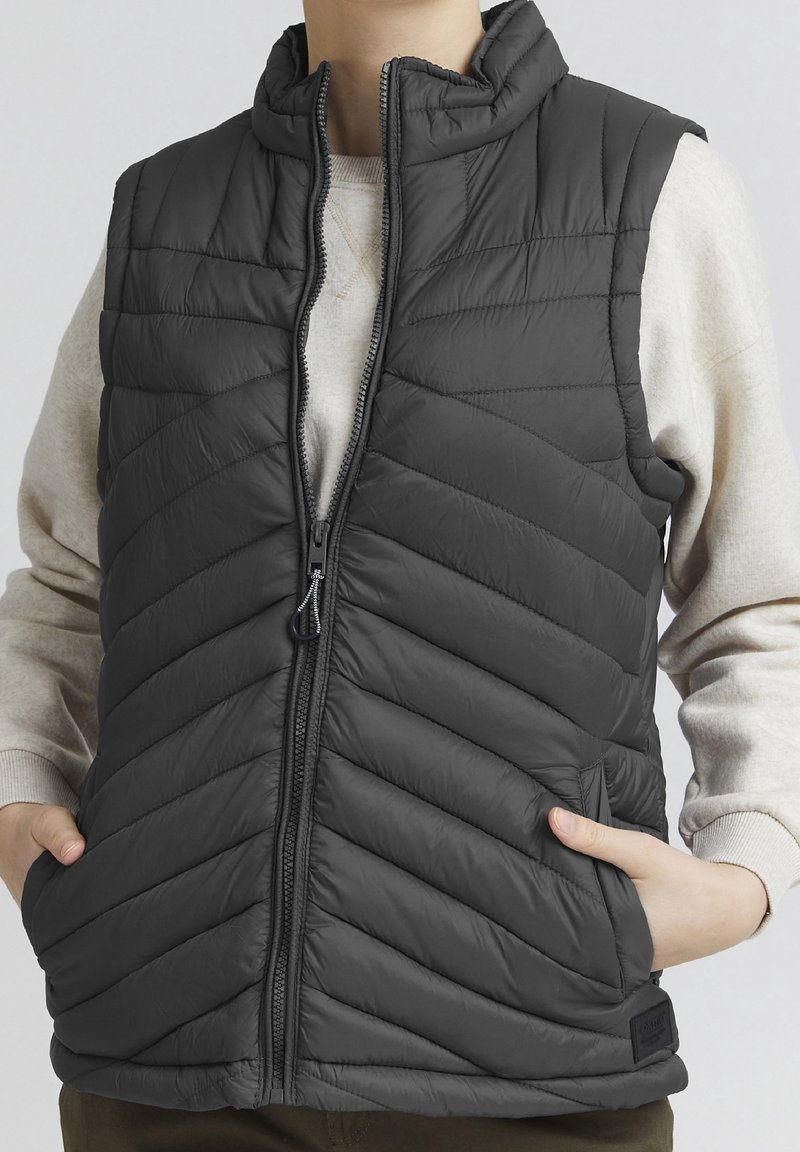 Personne portant une veste sans manches matelassée noire avec une fermeture éclair à l'avant et les mains dans les poches latérales, par-dessus une chemise beige clair à manches longues.