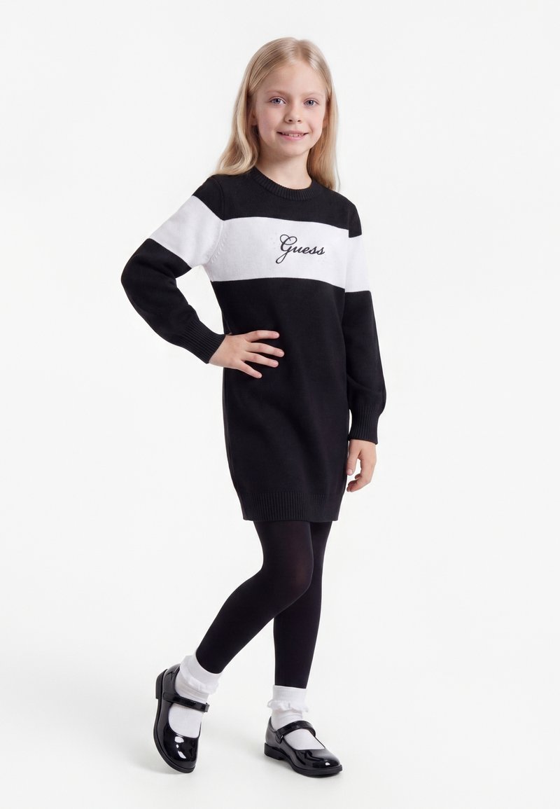 Jeune fille posant dans une robe-pull "Guess" noire et blanche, des collants noirs, des chaussettes blanches et des chaussures Mary Jane vernies noires sur fond blanc.