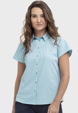 Junge Frau mit welligem braunem Haar, die ein hellblaues kurzärmeliges Hemd mit Knöpfen und dunkle Hosen vor einem schlichten Hintergrund trägt.