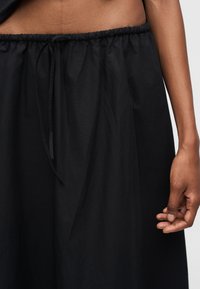 Μέση και χέρι ατόμου που φοράει φαρδιά μαύρη παντελόνα με ελαστική μέση και κορδόνι, με απλό φόντο.