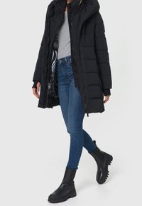 Sort puffermantel med hætte, featuring en lynlås og knaplukning, syede paneler og et mønstret indre for. Parret med blå jeans og sorte støvler.